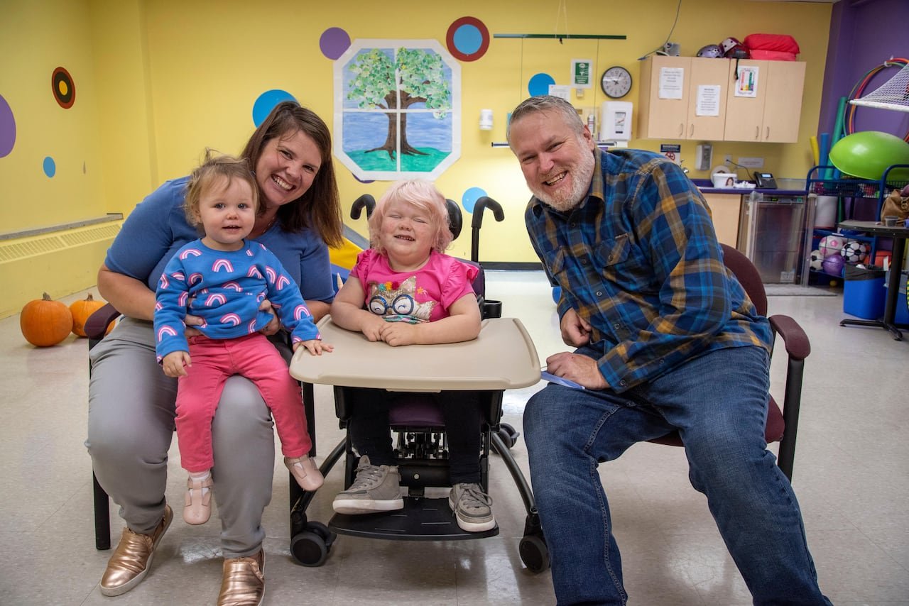 A smiling man and woman sit on either side of a smiling girl in a wheelchair. A smiling toddler sits on the lap of a woman.