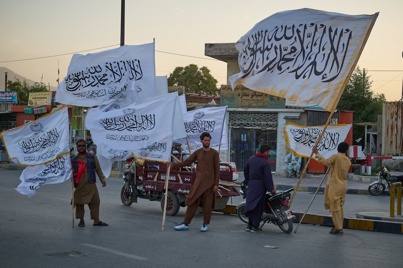 A group of men stand on the side of a paved road, all holding large flags with the Taliban sign on it.